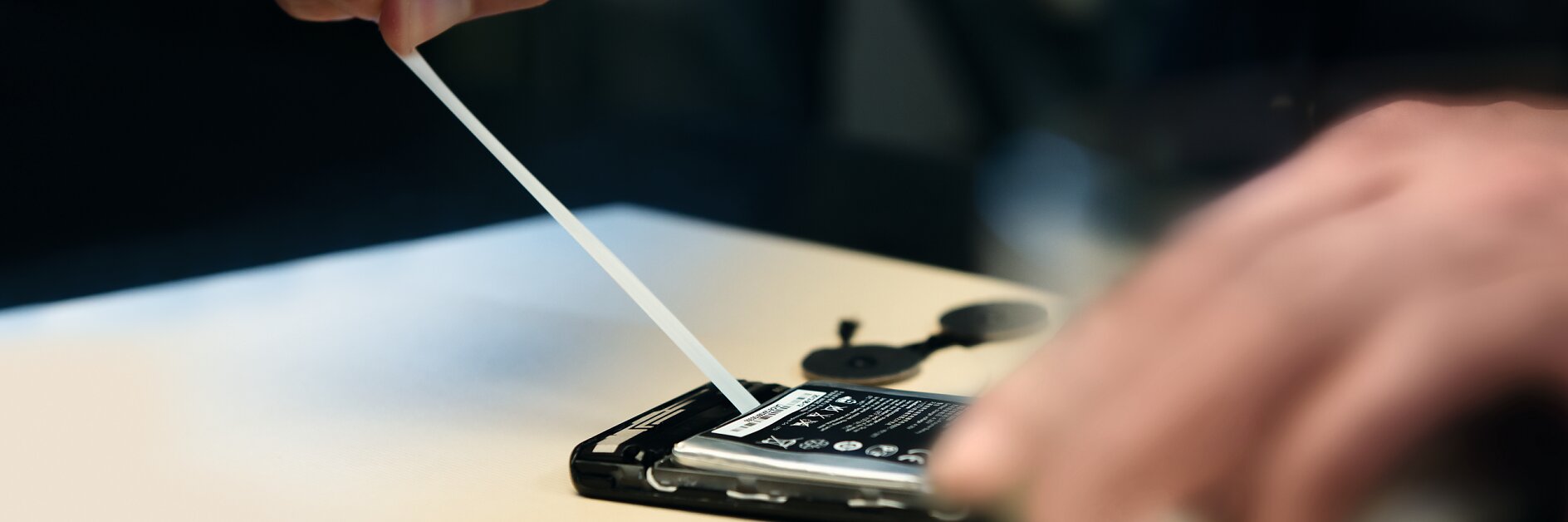 Close-up view of hands opening a smartphone to replace the battery, using a plastic tool. The phone is placed on a light-colored surface, and the battery is partially lifted from its compartment with tesa tape visible in the background.