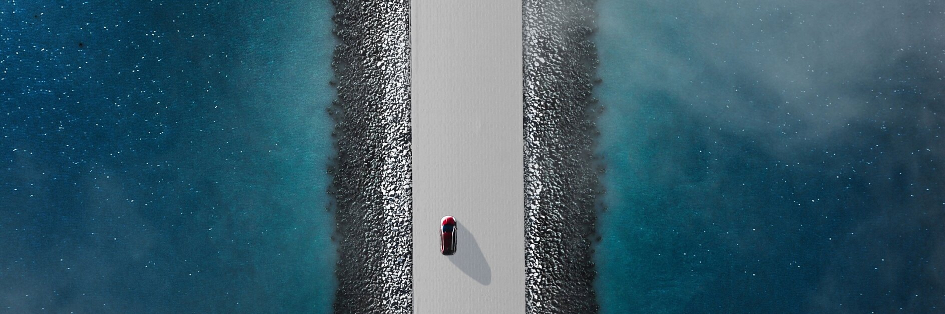 Aerial view of a red car driving on a narrow, straight road flanked by blue water on both sides. The road is elevated, with rocky edges lining the sides. The water appears calm with a slight mist or fog hovering above it.