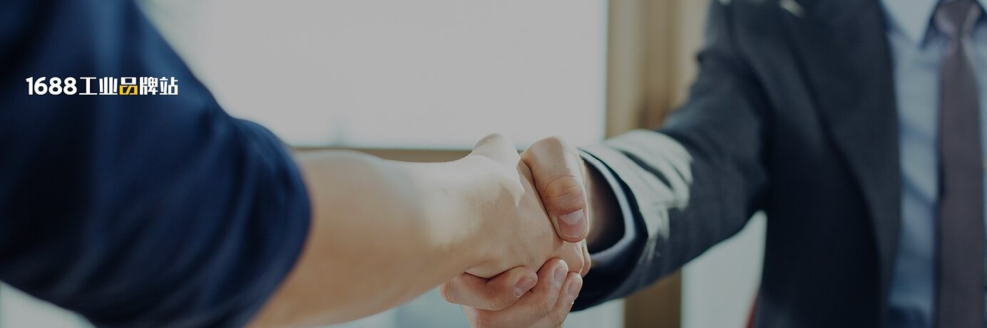 Two people are shaking hands over a desk with documents, charts, and a tablet on it. Both are dressed in business attire. The lighting suggests a daytime office setting.