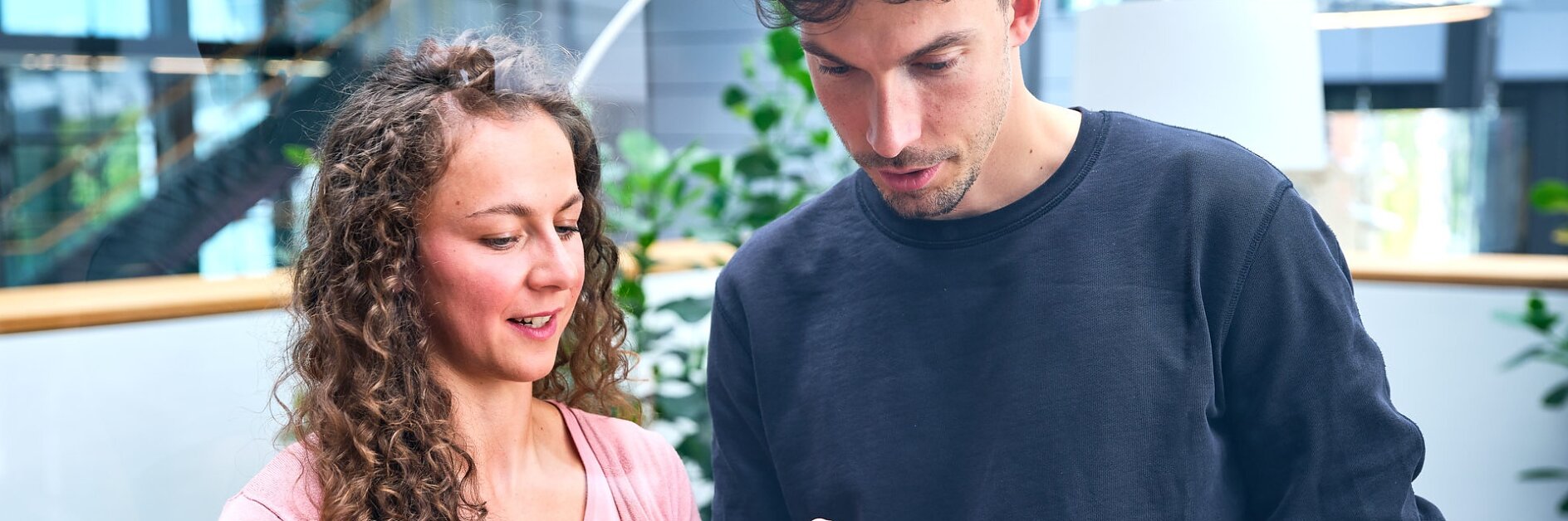 Two people are standing indoors, looking at a tablet. The woman on the left has curly hair and is wearing a pink sweater. The man on the right has short hair and is wearing a black sweatshirt. They appear to be discussing something on the screen, possibly related to tesa tape or its applications.