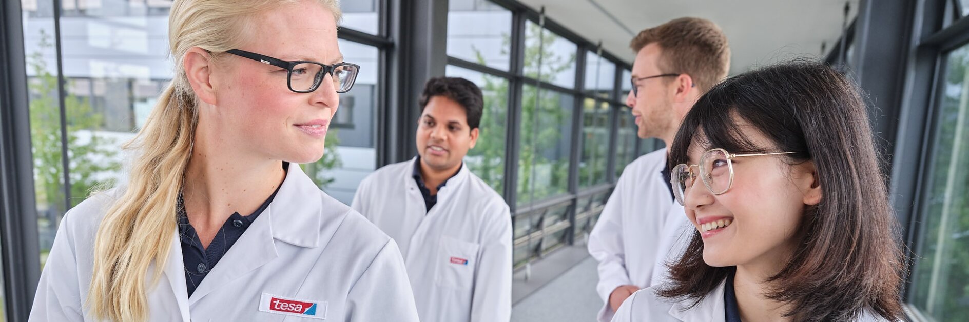 Four people in lab coats walk together along a corridor with large windows. Two individuals in the foreground, one with glasses and long blonde hair, and another with glasses and long dark hair, are looking at each other and smiling.