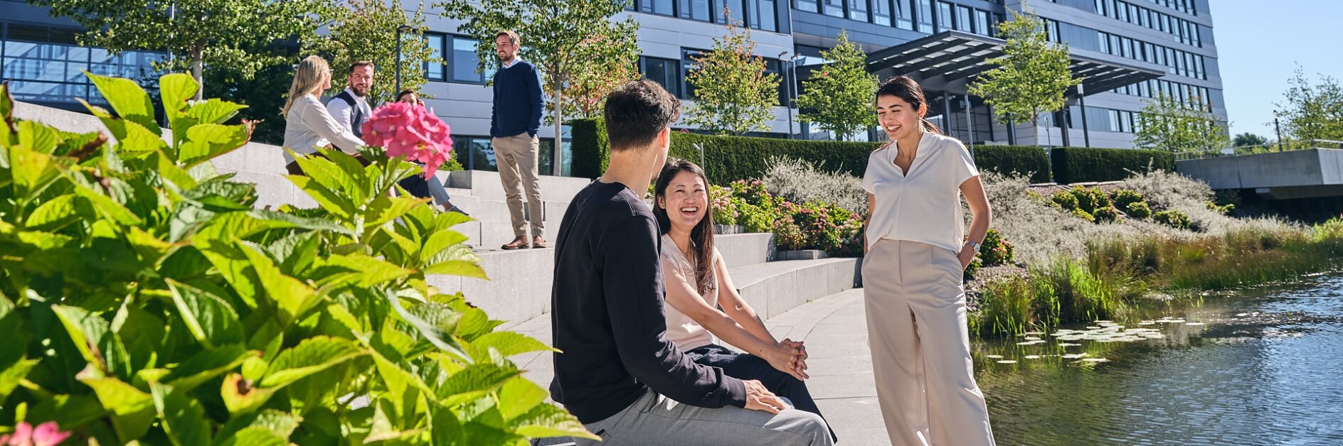 People are sitting and chatting on steps by a small pond with greenery in front of a modern multi-story building. The sky is clear and sunny, and the building has a sign that is partially visible with tesa tape.